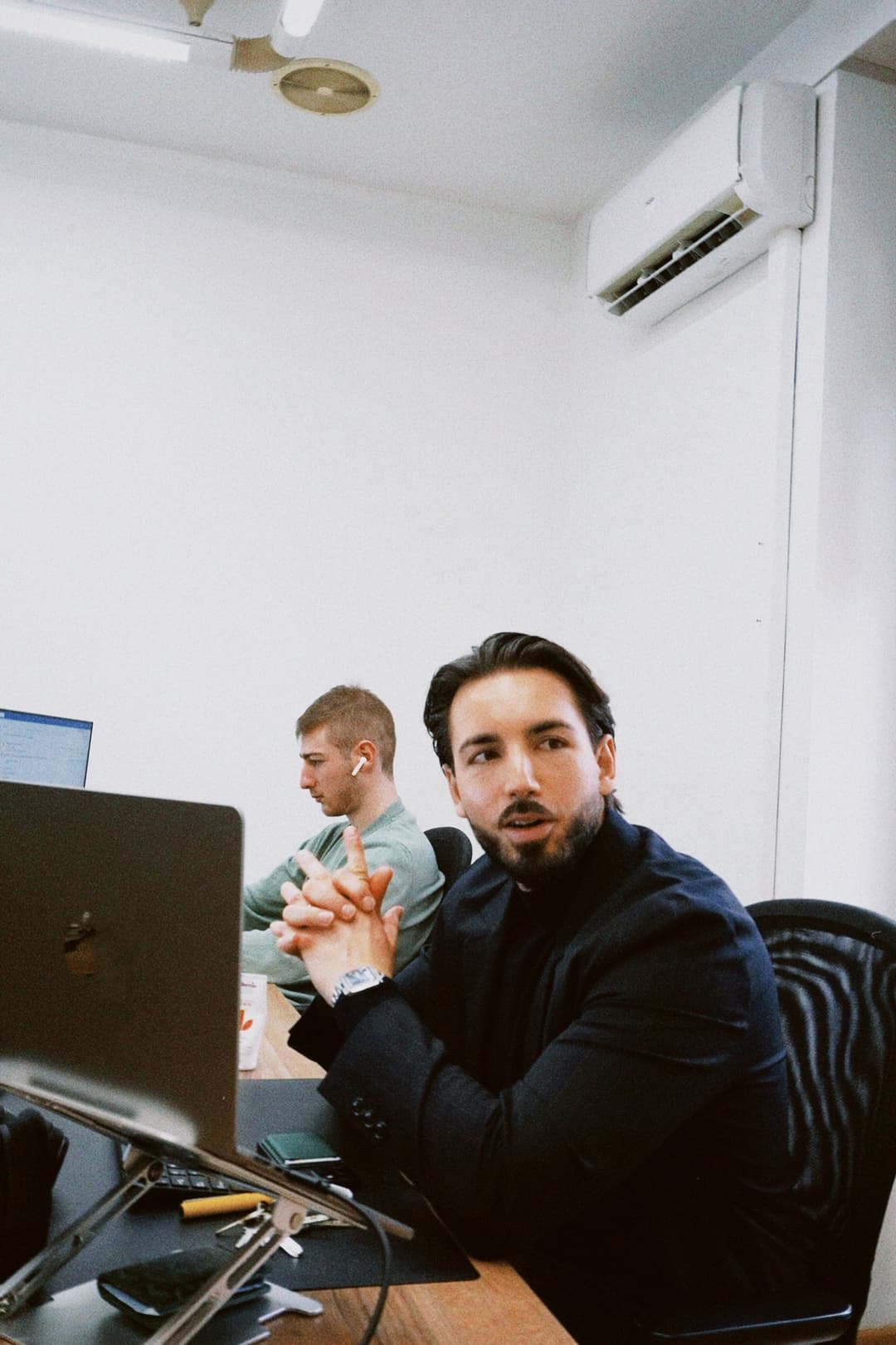 Two men seated at desks in a modern office environment, one in the foreground wearing a black blazer and gesturing with his hands while looking off-camera, the other in the background wearing a green shirt and white earbuds focused on a computer screen; visible elements include Apple laptops on adjustable stands, a wooden desk, black ergonomic chairs, a white wall-mounted air conditioning unit, and a ceiling-mounted light fixture with a circular vent; the scene conveys a collaborative or brainstorming work moment with natural lighting and a clean, minimal aesthetic.
