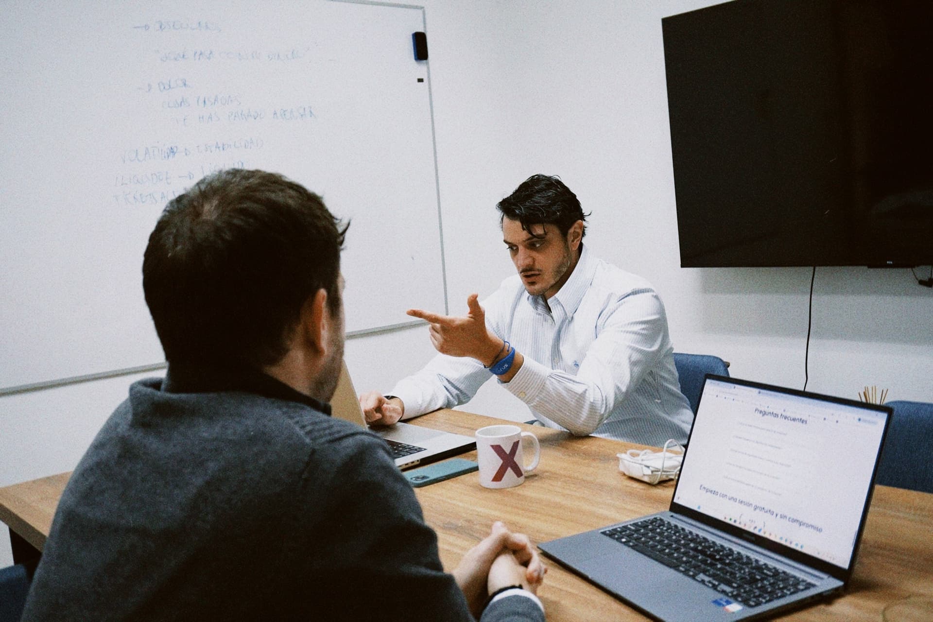 Two men engaged in a focused business meeting at a wooden table in a modern office setting; one man in a light blue striped shirt gestures emphatically while speaking, facing another man in a dark sweater seen from behind, with laptops open on the table displaying Spanish text including 'Preguntas frecuentes' and 'Empieza con una sesión gratuita y sin compromiso', a white mug with a red 'X' logo, a smartphone, and a whiteboard with handwritten Spanish notes visible in the background, alongside a mounted flat-screen monitor on the wall.