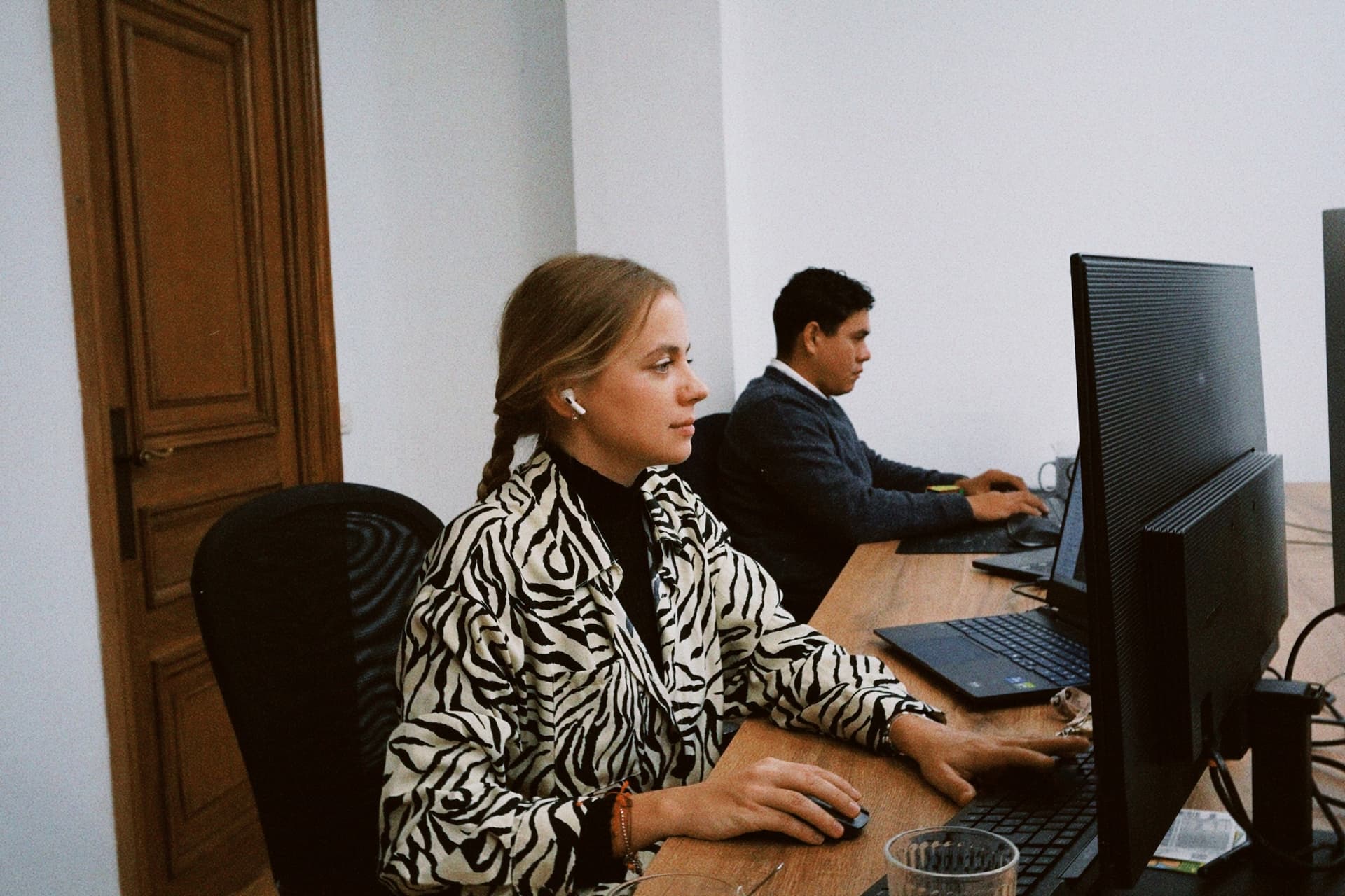 A woman with blonde hair styled in a side braid, wearing a black-and-white zebra-print jacket over a black top and white wireless earbuds, is seated at a wooden desk focused on a computer monitor, her right hand on a mouse and left hand near a keyboard; behind her, a man in a dark blue sweater is also seated at the same desk, working on a laptop with his hands on the keyboard; the setting includes a black office chair, a clear glass cup with pens, a second monitor, visible cables, and a wooden door with paneling to the left against a white wall, creating a professional yet casual office environment.